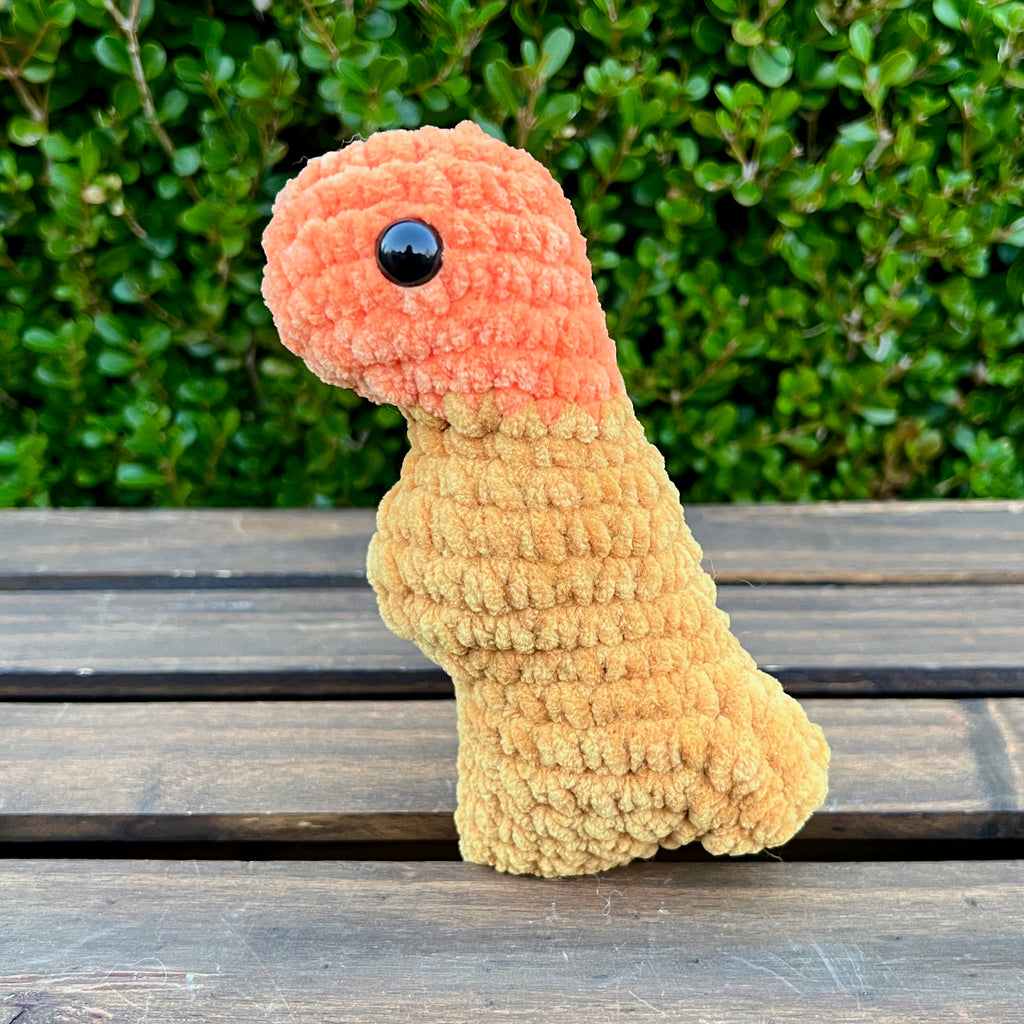 Crocheted dinosaur t-rex dino nugget with buffalo sauce dipped head on a wooden surface with green foliage in the background