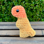 Crocheted dinosaur t-rex dino nugget with buffalo sauce dipped head on a wooden surface with green foliage in the background
