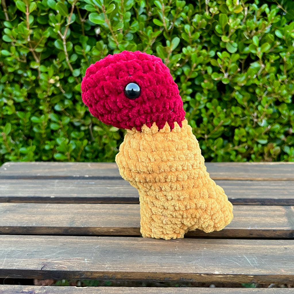 Crocheted dinosaur t-rex dino nugget with ketchup dipped head on a wooden surface with green foliage in the background