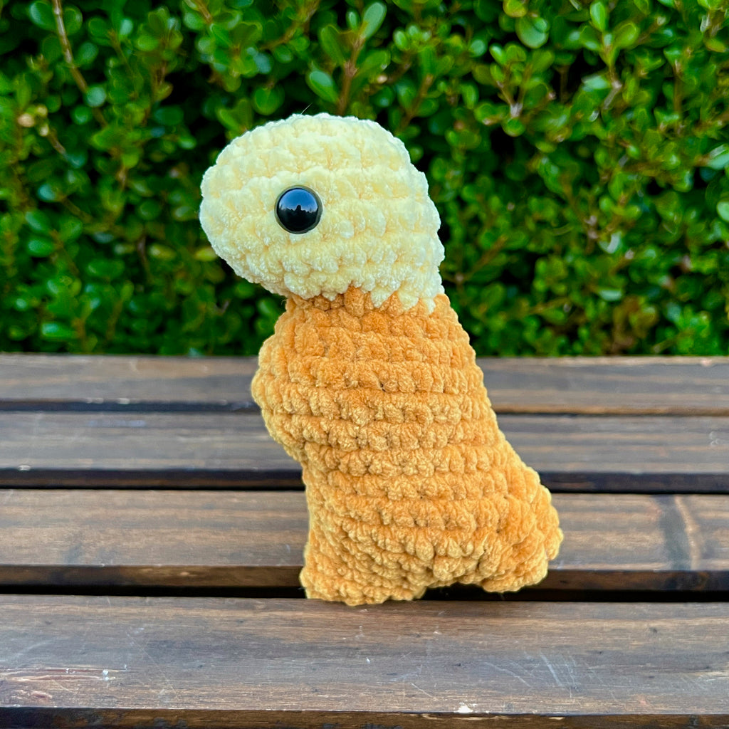 Crocheted dinosaur t-rex dino nugget with mustard dipped head on a wooden surface with green foliage in the background