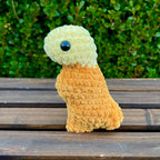 Crocheted dinosaur t-rex dino nugget with mustard dipped head on a wooden surface with green foliage in the background