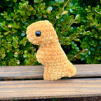 Crocheted t-rex dino nugget on a wooden bench with green foliage in the background